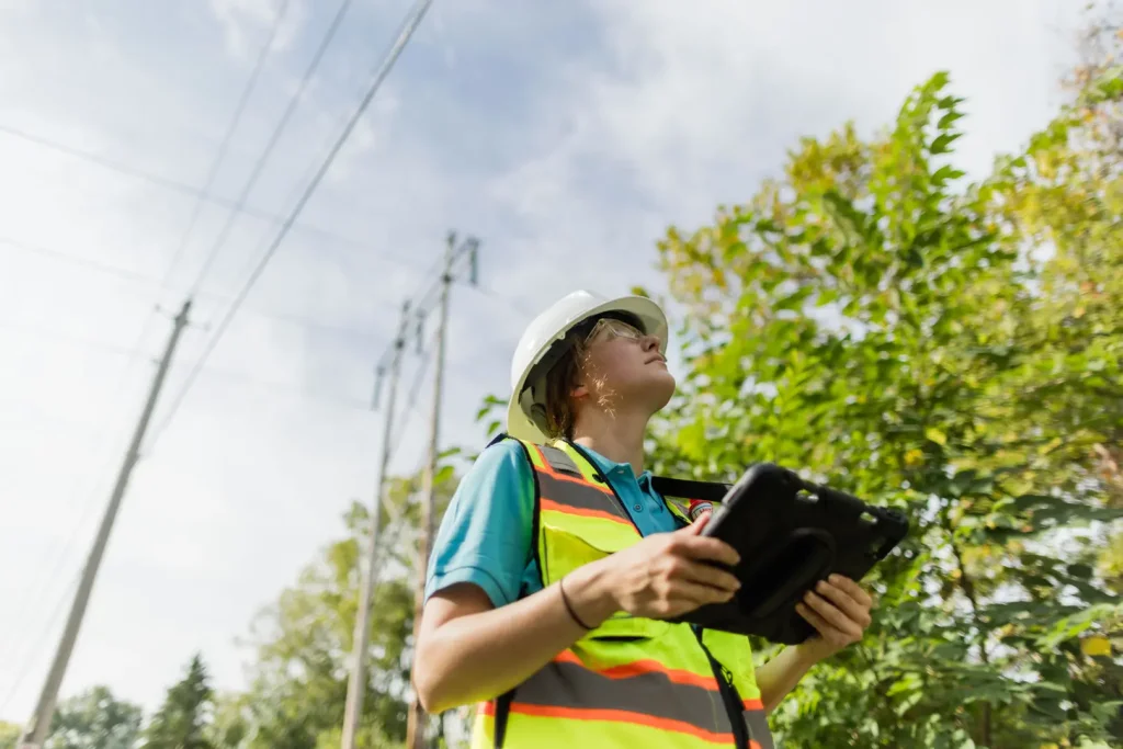 A worker wearing a hard hat and safety vest inspects power lines near trees
